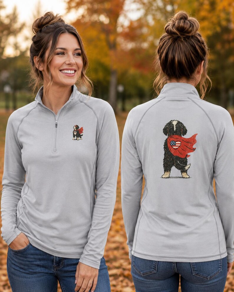 K&ouml;nnte beinhalten: Ein hellgrauer Langarm-Pullover mit kurzem Rei&szlig;verschluss, der auf der linken Brust ein kleines Motiv eines Berner Sennenhundes mit rotem Umhang und auf der R&uuml;ckseite ein gr&ouml;&szlig;eres, passendes Motiv des Hundes mit einer herzf&ouml;rmigen US-Flagge zeigt.