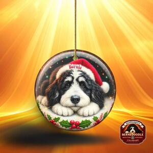 May include: Round Christmas ornament featuring a Bernedoodle puppy wearing a Santa hat with the name "Bernie". The ornament has a gold string for hanging and is decorated with holly and berries. The background is a warm, golden color.