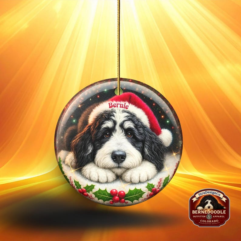 May include: Round Christmas ornament featuring a Bernedoodle puppy wearing a Santa hat with the name "Bernie". The ornament has a gold string for hanging and is decorated with holly and berries. The background is a warm, golden color.