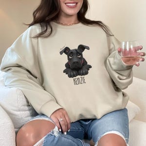 Puede incluir: Sudadera beige con un gr&aacute;fico de cachorro negro y el nombre "KENZIE" impreso debajo. El cachorro tiene un collar rojo y una expresi&oacute;n triste. La sudadera es de color neutro y tiene mangas largas.
