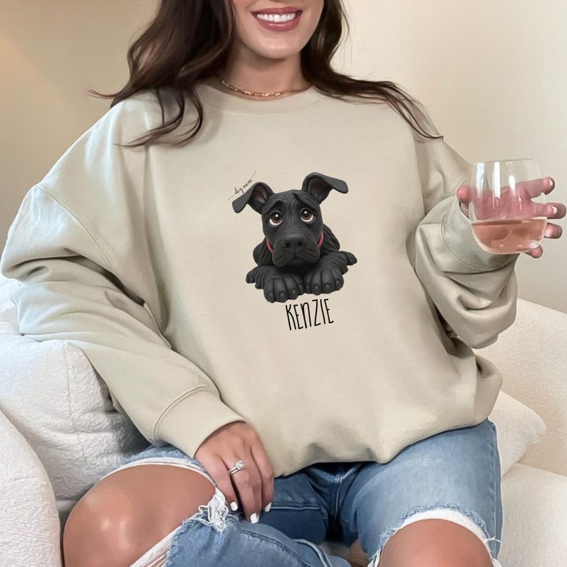 Puede incluir: Sudadera beige con un gr&aacute;fico de cachorro negro y el nombre "KENZIE" impreso debajo. El cachorro tiene un collar rojo y una expresi&oacute;n triste. La sudadera es de color neutro y tiene mangas largas.