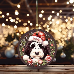 May include: A round Christmas ornament featuring a black and white dog wearing a Santa hat with the name "Bernie". The ornament is decorated with a wreath of greenery, red flowers, and snowflakes, and two red and white ornaments.