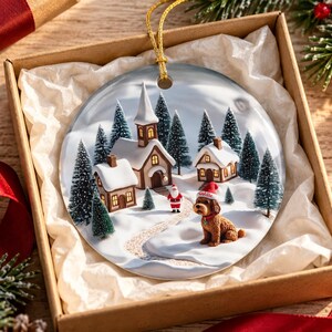 May include: A round ceramic Christmas ornament featuring a snowy village scene with a church, houses, a small Santa Claus figure, and a brown dog wearing a red Santa hat with the name Eddie printed on it, hanging by a gold cord.