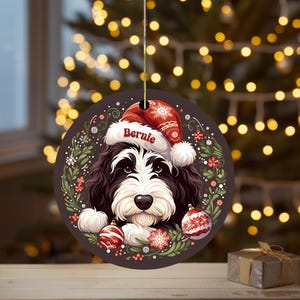 May include: Round Christmas ornament featuring a dog wearing a Santa hat with the name "Bernie". The ornament has a dark background with a wreath of red and white flowers and ornaments. A small gift box is in the foreground.