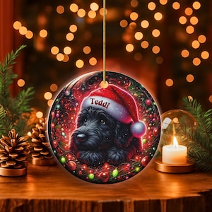 May include: A round Christmas ornament featuring a black dog wearing a Santa hat with the name "Teddi" on it. The ornament has a festive design with red and green accents, and is hanging from a gold string.