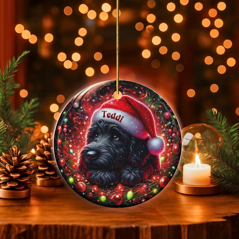 May include: A round Christmas ornament featuring a black dog wearing a Santa hat with the name "Teddi" on it. The ornament has a festive design with red and green accents, and is hanging from a gold string.