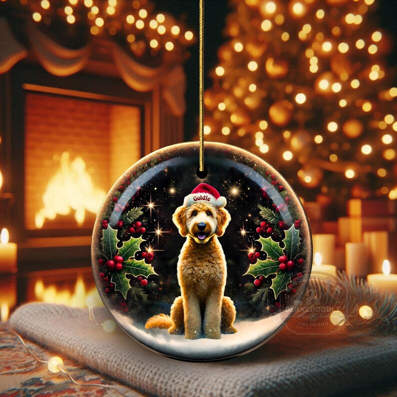 May include: A round Christmas ornament featuring a golden doodle dog wearing a Santa hat. The ornament has a festive design with holly, berries, and a snowy base. The background includes a fireplace and Christmas tree.