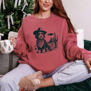 May include: A maroon sweatshirt featuring a graphic of a dog wearing a cowboy hat and bandana, with a desert landscape and a UFO. The dog is the central focus, set against a backdrop of cacti and a flying saucer.