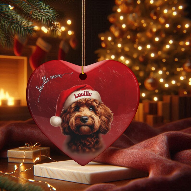 May include: Red heart-shaped Christmas ornament featuring a brown Doodle dog wearing a Santa hat. The ornament has the name "Lucille" and the words "doodle mom" printed on it. The ornament is hanging in front of a Christmas tree.