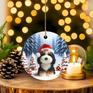 Bernedoodle claymation style ornament made of ceramic, hanging by a gold string.  Winter scene featuring a cute, personalized doodle dog with personalized name on santa hat.  Perfect gift for Bernedoodle Dog Lovers and Pet Enthusiasts.