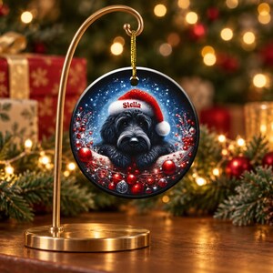 May include: A round ceramic Christmas ornament hanging from a gold metal stand. The ornament features a black dog wearing a red Santa hat with the name Stella printed on the brim, surrounded by red berries and ornaments against a dark background.