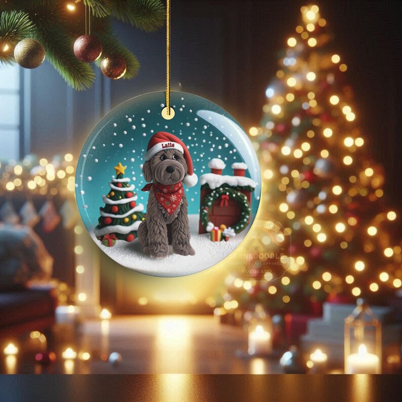 May include: A clear glass Christmas ornament featuring a brown dog wearing a Santa hat and a red bandana. The ornament has a snowy scene with a Christmas tree and a chimney. The dog's name, "Lalle", is on the hat.