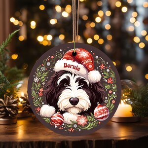 May include: A round Christmas ornament featuring a black and white dog wearing a Santa hat with the name "Bernie". The ornament is decorated with a wreath of greenery, red flowers, and two red and white ornaments. A brown string is attached for hanging.