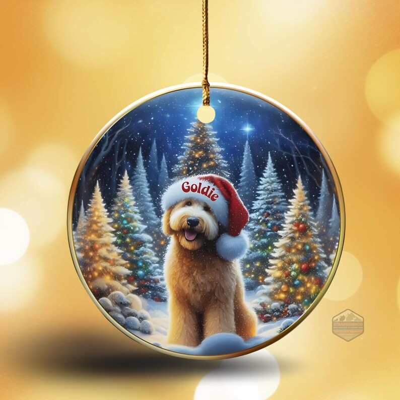 May include: A round Christmas ornament featuring a golden doodle wearing a Santa hat with the name "Goldie" on it. The background shows a snowy forest with Christmas trees. The ornament has a gold-colored border and a gold string for hanging.