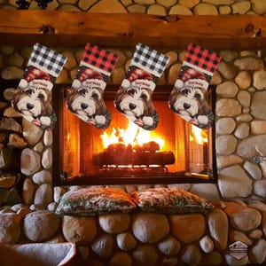 A festive Christmas stocking featuring a whimsical illustrated doodle dog wearing a red Santa hat, surrounded by holiday ornaments and greenery in a warm, detailed Christmas art style. The top of the stocking has a classic red and black buffalo-plaid cuff, and the stocking hangs from a white mantel decorated with evergreen garland and gold ornaments. The stocking design is personalized with your dog’s name, making it a charming and custom holiday décor piece for dog lovers.