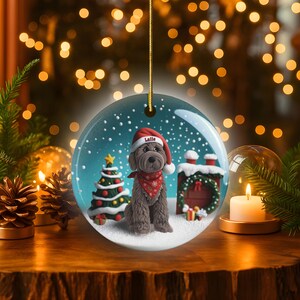 May include: A round Christmas ornament featuring a dog wearing a Santa hat and a red bandana. The ornament has a Christmas tree and a house with a wreath. The dog's hat says "Latte". The background is a snowy scene.
