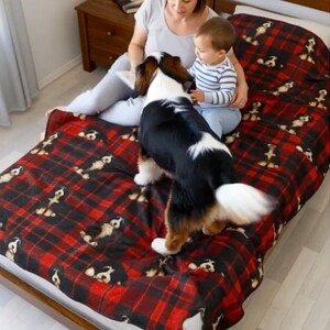 Peut inclure: Une couverture douce et moelleuse orn&eacute;e d'un motif &agrave; carreaux rouge et noir avec une impression r&eacute;p&eacute;titive de petites illustrations de chiens. La couverture est pos&eacute;e sur un lit, mettant en valeur la texture du tissu et les graphismes canins.