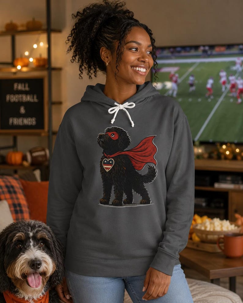 Pu&ograve; includere: Una felpa con cappuccio grigio antracite con la grafica di un cane nero che indossa un mantello da supereroe rosso, una maschera rossa e un emblema a forma di cuore sul petto con strisce rosse, bianche e blu. La felpa ha cordini bianchi.