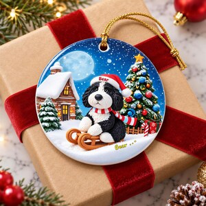 A ceramic ornament featuring a black and white dog wearing a Santa hat, sitting on a sled and looking at the camera, with a snowy village scene and a full moon in the background.