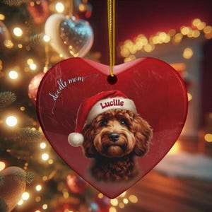 May include: Heart-shaped Christmas ornament with a red background. The ornament features a portrait of a brown dog wearing a Santa hat with the name "Lucille" on it. The words "doodle mom" are written on the ornament.