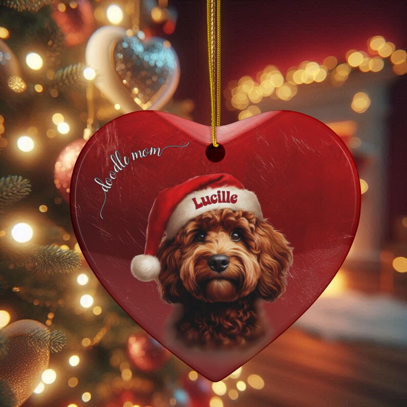 May include: Heart-shaped Christmas ornament with a red background. The ornament features a portrait of a brown dog wearing a Santa hat with the name "Lucille" on it. The words "doodle mom" are written on the ornament.