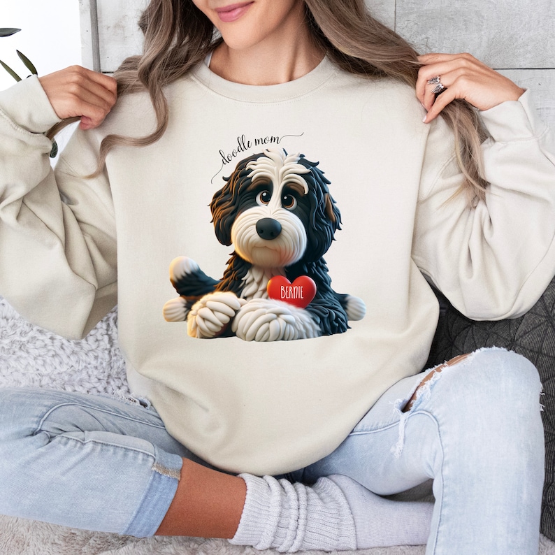 A woman wearing a Bernedoodle sweatshirt with cute design on the front and the back.  The front features a bernedoodle dog in claymation design holding a heart with his name, across the top of his head the words, "Doodle Mom".