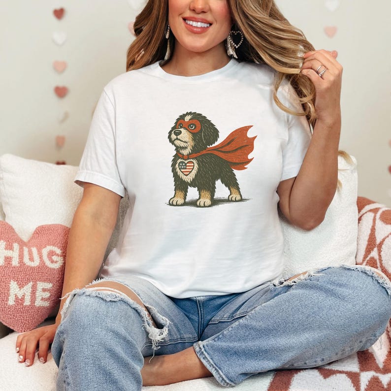 May include: White t-shirt featuring a cartoon dog dressed as a superhero. The dog has a red mask, cape, and a heart-shaped emblem with an American flag design. The dog is standing on a white background.