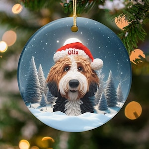 Könnte beinhalten: Runder Weihnachtsschmuck mit einem Hund, der einen Weihnachtsmannhut mit dem Namen "Otis" trägt. Der Hund hat braunes, weißes und schwarzes Fell, vor einer verschneiten Winterlandschaft mit Kiefern. Ein goldener Faden ist zum Aufhängen befestigt.