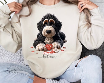 May include: A cream-colored crewneck sweatshirt featuring a graphic of a black and white doodle dog lying down with its paws on a red heart that says Love. Below the dog, the text doodle mom is written in a cursive script with small red hearts.
