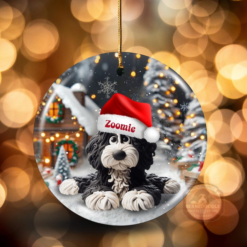 May include: A round Christmas ornament featuring a black and white Bernedoodle dog wearing a red Santa hat with the name "Zoomie". The ornament has a winter scene background with a house, trees, and snowflakes. A gold string is attached for hanging.