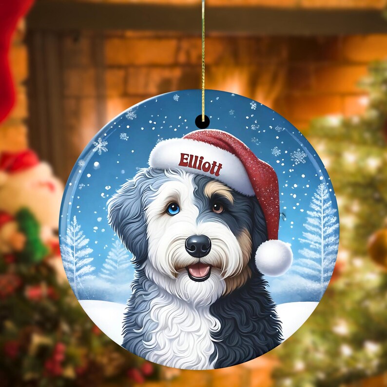 May include: A round ceramic Christmas ornament featuring a fluffy dog with black, white, and tan fur, wearing a red Santa hat with the name Elliott printed in white. The background shows a snowy winter scene with blue tones and pine trees.