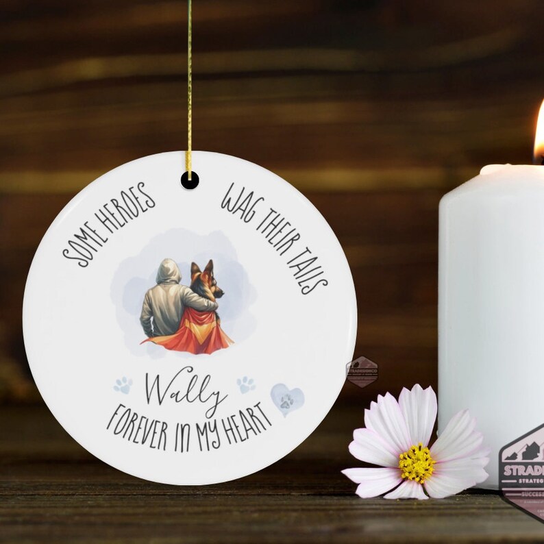 May include: White ceramic ornament with a gold string, featuring a watercolor illustration of a person and a German Shepherd dog. Text reads "Some Heroes Wag Their Tails" and "Wally Forever in My Heart."
