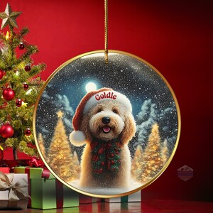 May include: A round Christmas ornament featuring a fluffy, light brown dog wearing a Santa hat with the name "Goldie" and a festive green and red garland. The background shows a snowy winter scene with Christmas trees and a full moon. The ornament hangs from a gold string.