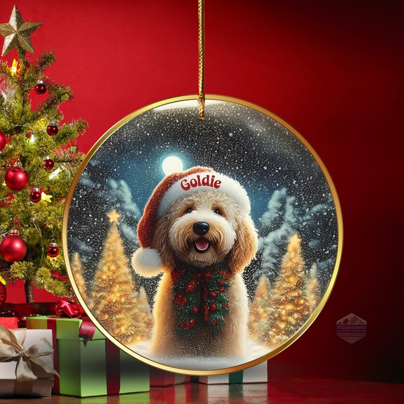May include: A round Christmas ornament featuring a fluffy, light brown dog wearing a Santa hat with the name "Goldie" and a festive green and red garland. The background shows a snowy winter scene with Christmas trees and a full moon. The ornament hangs from a gold string.