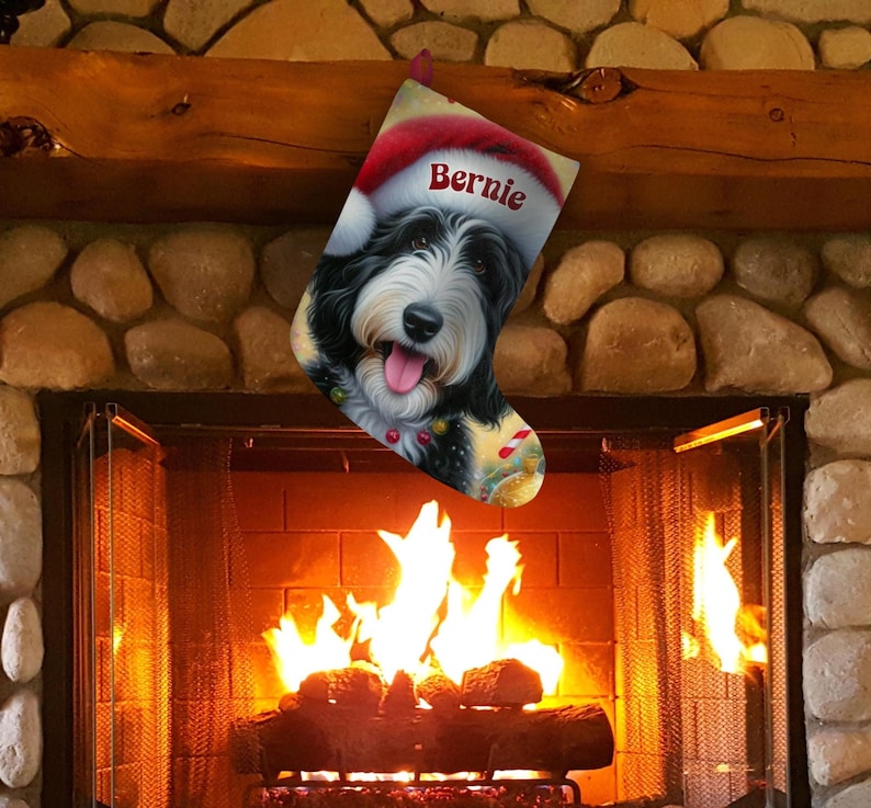 Op de afbeelding: Een kerstsok met een zwart-witte hond met een kerstmuts met de naam "Bernie" erop. De sok hangt boven een open haard met een brandend vuur.