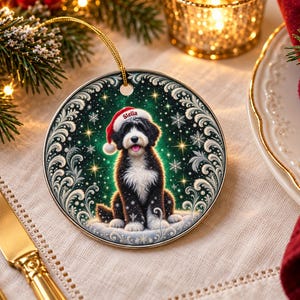 May include: A round ceramic Christmas ornament featuring a black and white dog wearing a red Santa hat with the name Stella printed on the brim. The dog sits against a dark green background with white snowflakes, gold stars, and a silver filigree border.