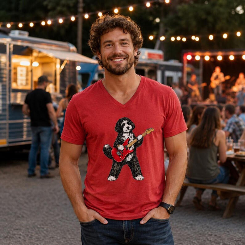 May include: A red V-neck t-shirt featuring a graphic illustration of a black and white doodle dog standing on its hind legs while playing a red electric guitar. The shirt is made of a soft, heathered fabric and has short sleeves.