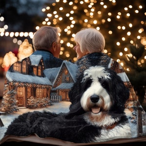 May include: A cozy blanket featuring a winter scene with a snow-covered house and a black and white dog. The blanket is draped over a couple, with a blurred background of Christmas lights.