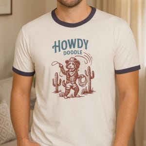 May include: Cream-colored ringer t-shirt with a navy blue trim featuring a graphic of a cowboy doodle dog roping a lasso. The text "HOWDY DOODLE" is above the dog illustration, with two cacti on either side.