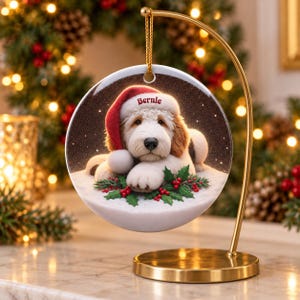 May include: A round ceramic Christmas ornament hanging from a gold metal stand. The ornament features a fluffy white and tan dog wearing a red Santa hat with the name Bernie printed on it, resting on green holly leaves and red berries.