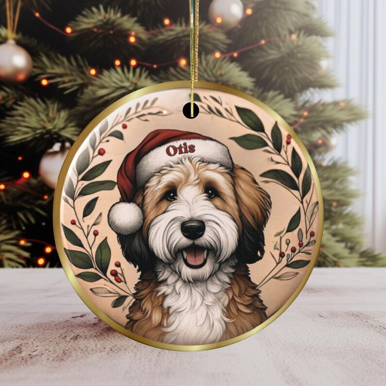 Puede incluir: Un adorno redondo de cer&aacute;mica con borde dorado que presenta la ilustraci&oacute;n de un perro marr&oacute;n y blanco con un gorro de Pap&aacute; Noel rojo con el nombre Otis impreso en el borde, rodeado por una corona decorativa de hojas verdes y bayas rojas.