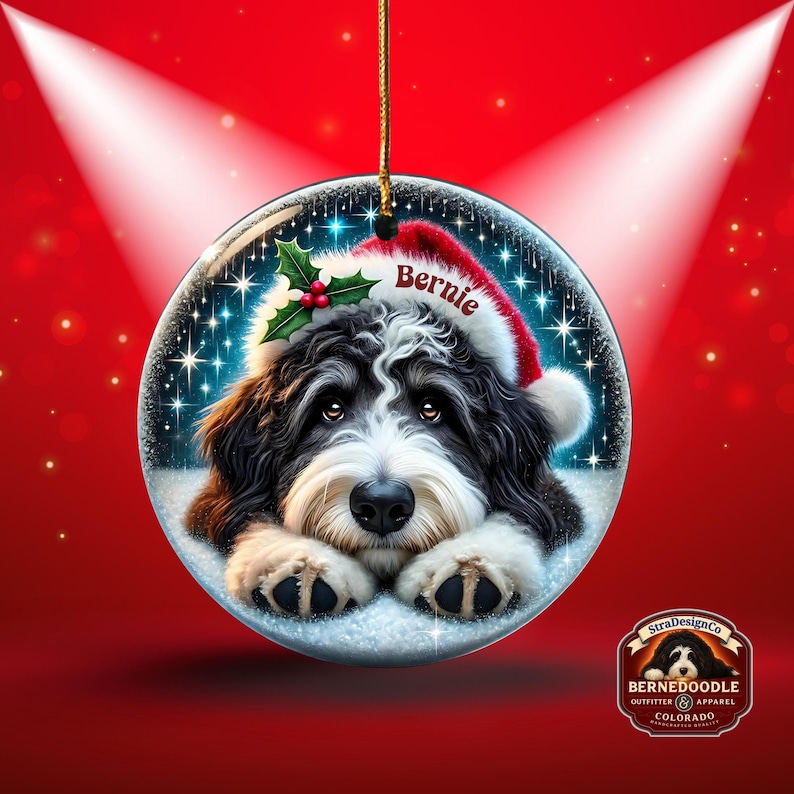 May include: A round Christmas ornament featuring a black and white Bernedoodle dog wearing a Santa hat with the name "Bernie". The ornament has a gold string for hanging and is set against a red background with spotlights.