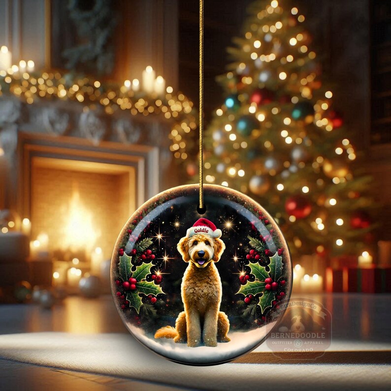May include: A round Christmas ornament featuring a golden doodle dog wearing a Santa hat. The ornament has a black background with stars, holly, and berries. The dog's name, "Goldie", is on the hat. The ornament is hanging in front of a Christmas tree.