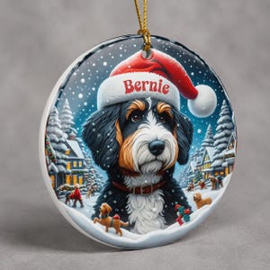 A decorative ornament featuring a Bernese mountain dog wearing a Santa hat, surrounded by a snowy winter landscape with houses, trees, and people.