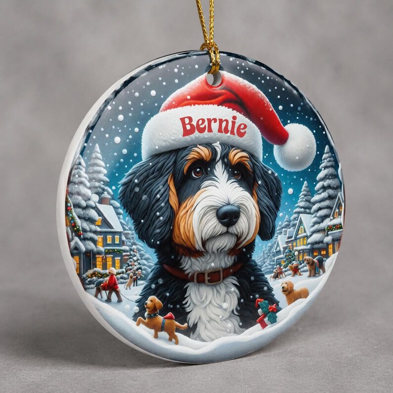 A decorative ornament featuring a Bernese mountain dog wearing a Santa hat, surrounded by a snowy winter landscape with houses, trees, and people.