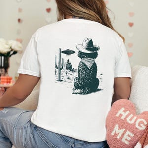 May include: White t-shirt featuring a black and white graphic of a dog wearing a cowboy hat and bandana, with a desert landscape and UFO in the background. The design is centered on the back of the shirt.