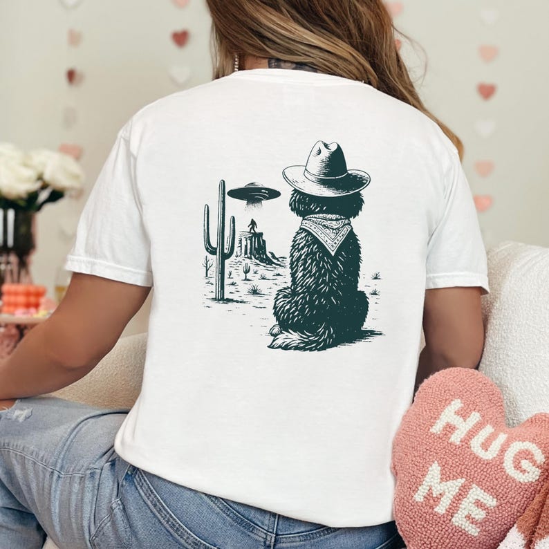 May include: White t-shirt featuring a black and white graphic of a dog wearing a cowboy hat and bandana, with a desert landscape and UFO in the background. The design is centered on the back of the shirt.