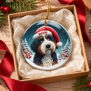 May include: A round ceramic Christmas ornament featuring a Bernese Mountain Dog wearing a red Santa hat with the name Bernie printed on the brim. The dog is set against a snowy winter landscape with pine trees and snowmen in the background.