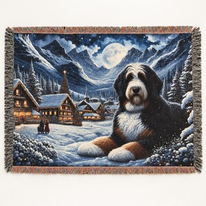 Puede incluir: Una manta tejida que muestra a un perro de monta&ntilde;a de Berna descansando frente a un pueblo invernal nevado de noche, con casas iluminadas, monta&ntilde;as cubiertas de nieve, una luna llena y bordes con flecos.