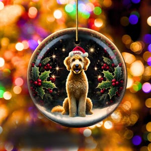 May include: A round Christmas ornament featuring a golden doodle dog wearing a Santa hat with the name "Goldie". The ornament has a festive design with holly, berries, and a snowy base. A gold string is attached for hanging.